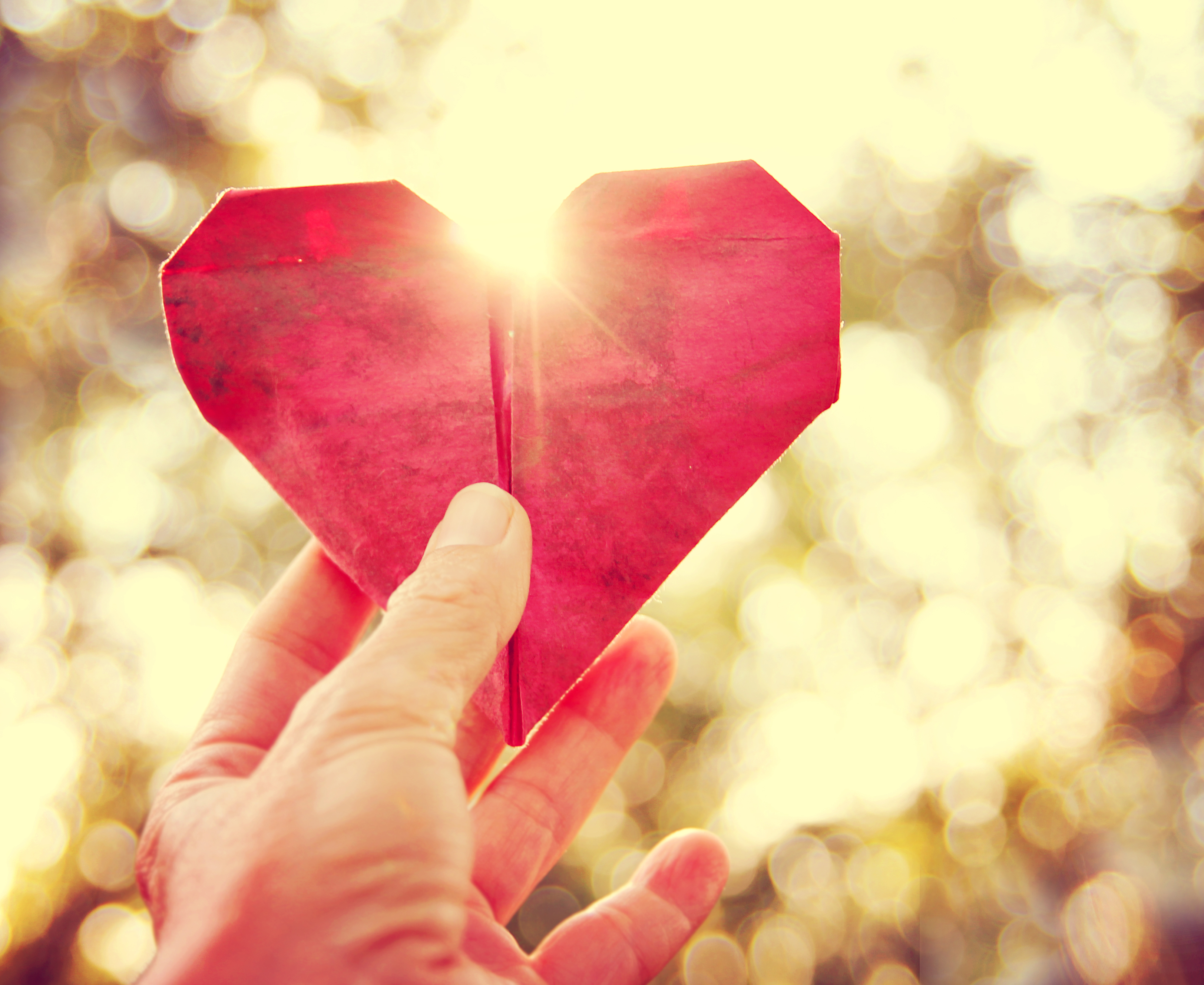 a hand holding an origami paper heart up to the sun during sunse